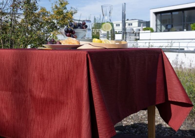 Bordeauxfarbene Jacquard-Tischdecke mit feiner Struktur, dekoriert mit Obst und Getränken auf einem Tisch.