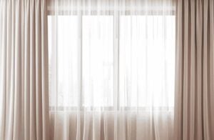 Das Bild zeigt eine große Fensterfront mit weißen Vorhängen und seitlichen beige Gardinen.